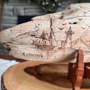 May include: A carved wooden sculpture of a nautical scene, featuring sailboats, a lighthouse, and flying birds. The sculpture is mounted on a wooden stand and has the name "A. LOWTHER" carved into the wood.