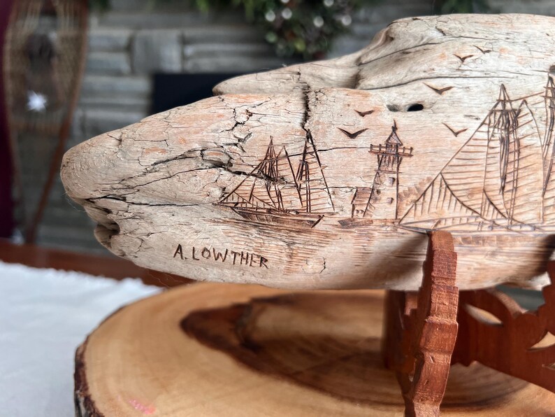 May include: A carved wooden sculpture of a nautical scene, featuring sailboats, a lighthouse, and flying birds. The sculpture is mounted on a wooden stand and has the name "A. LOWTHER" carved into the wood.