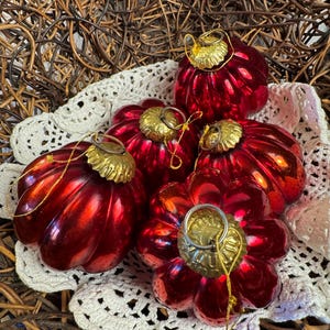 May include: Five shiny, deep red glass ornaments with a ribbed, pumpkin-like shape. Each ornament features a metallic gold-colored cap at the top with a small loop for hanging. The ornaments are arranged in a cluster on a white crocheted doily.