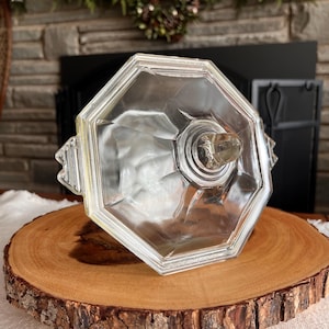 Könnte beinhalten: Eine klare, achteckige Bonbonniere aus Glas mit Deckel, die auf einer runden Holzscheibe ruht. Die Schale hat ein facettiertes Design und einen kleinen Knopf oben. Die Holzscheibe hat einen natürlichen Rindenrand.