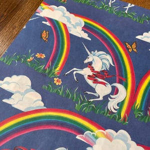 May include: Blue wrapping paper featuring a pattern of white unicorns, rainbows, clouds, butterflies, and flowers. The unicorns have flowing manes and red ribbons. The rainbows are in bright colours.