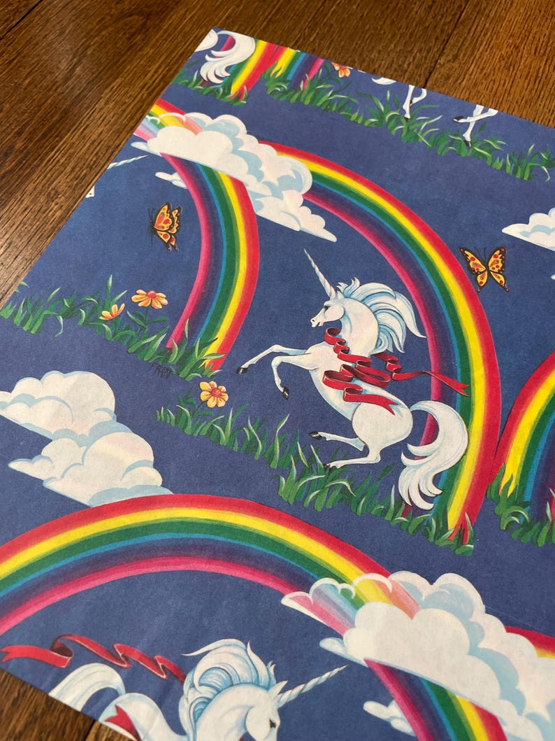 May include: Blue wrapping paper featuring a pattern of white unicorns, rainbows, clouds, butterflies, and flowers. The unicorns have flowing manes and red ribbons. The rainbows are in bright colours.