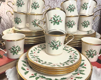 Puede incluir: Un juego de vajilla de porcelana color crema con un patrón festivo de acebo y bayas con bordes dorados. El conjunto incluye varios platos apilados y varias tazas a juego, cada una decorada con hojas de acebo verdes y bayas rojas.