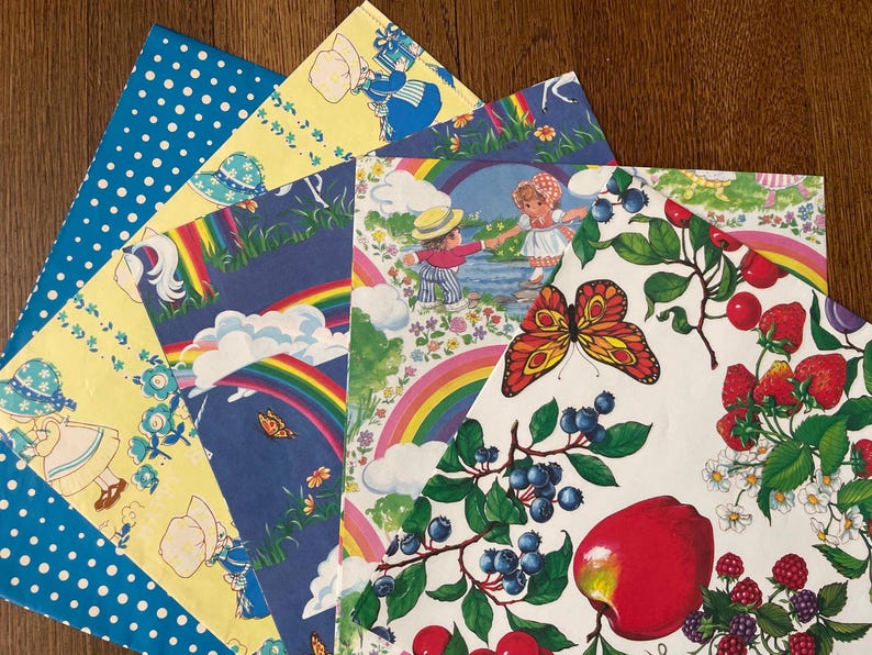 May include: A selection of patterned papers featuring various designs. The papers display illustrations of children, rainbows, butterflies, and fruit. The papers are arranged in a fan shape, showcasing the variety of illustrations and colours.