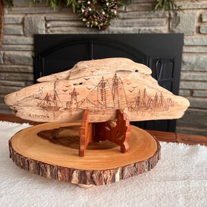 May include: A wooden sculpture of a coastal scene, featuring sailboats, a lighthouse, and flying birds, is displayed on a wooden stand. The sculpture is mounted on a round, natural wood slice base. The artist's name, A. Lowther, is visible.