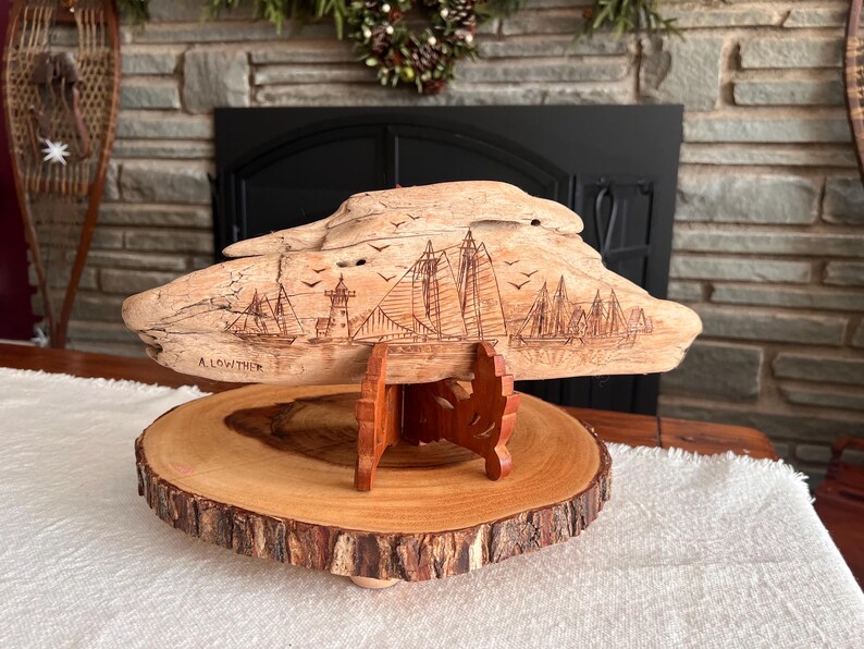 May include: A wooden sculpture of a coastal scene, featuring sailboats, a lighthouse, and flying birds, is displayed on a wooden stand. The sculpture is mounted on a round, natural wood slice base. The artist's name, A. Lowther, is visible.
