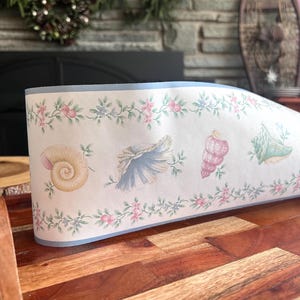 May include: A roll of wallpaper with a seashell design. The wallpaper has a light background with a blue border and a repeating pattern of seashells in various colours, including pink, blue, and yellow, interspersed with floral elements.