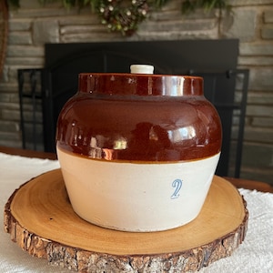 Puede incluir: Un tarro de cerámica bicolor marrón y blanco con tapa, sobre una rodaja de madera. El tarro tiene un acabado brillante y el número "2" está impreso en azul en la sección blanca. El fondo incluye una chimenea y vegetación.