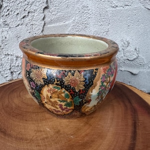 May include: A round, ceramic planter with a wide opening. The planter features a black background with colorful floral and bird designs. The rim is trimmed with gold and red accents. The planter sits on a wooden surface.