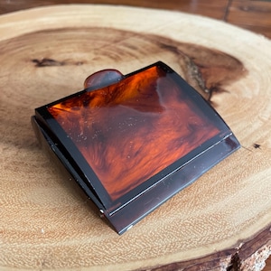 May include: A vintage, rectangular compact with a tortoiseshell pattern. The compact features a dark brown frame and a matching clasp. The surface has a rich, swirling pattern of amber and brown hues, suggesting a classic, retro design.