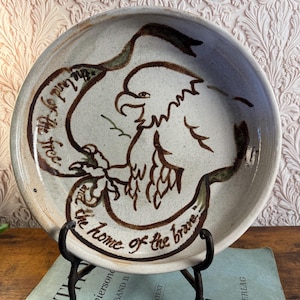 May include: A round ceramic plate with a speckled off-white glaze featuring a brown hand-painted eagle and a ribbon banner. The text on the banner reads, "the land of the free and the home of the brave." The plate is displayed on a metal stand.