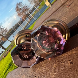 May include: Two octagonal door knobs made of translucent purple glass with faceted edges. Each knob features a circular metallic base with a brass-toned finish and visible screw holes for mounting, reflecting light through the glass surfaces.