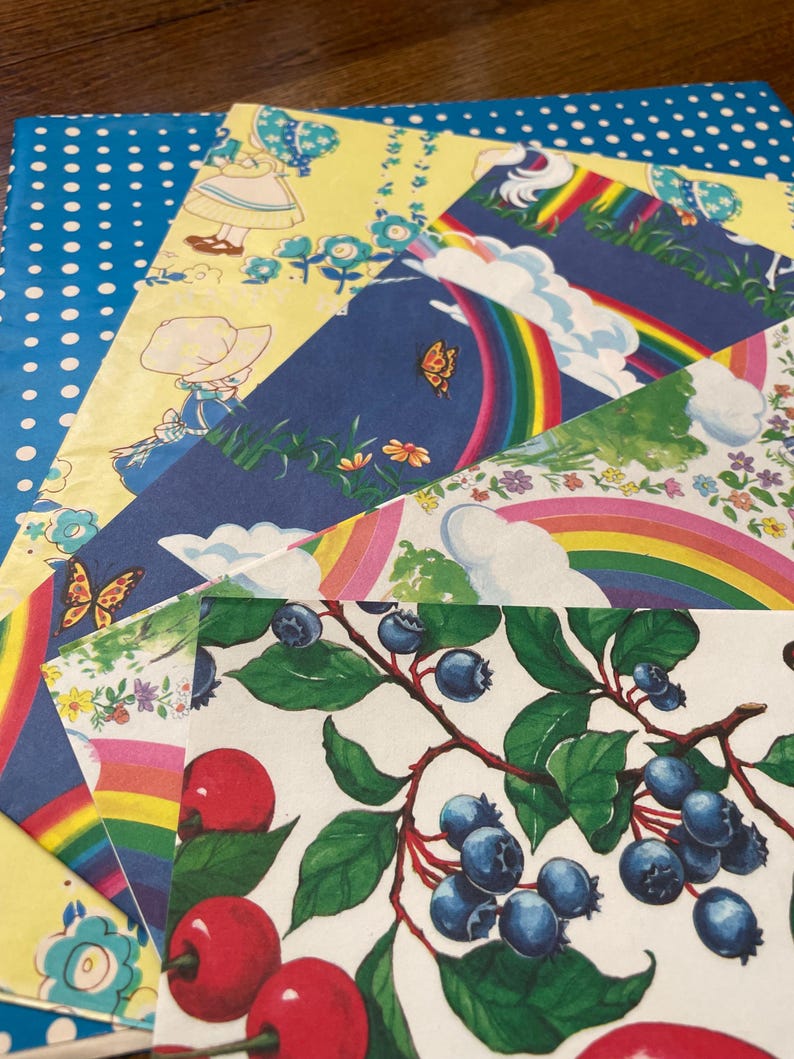 May include: A selection of colourful vintage paper sheets with diverse patterns. Designs include a blue polka dot pattern, a yellow floral pattern with a girl, a rainbow with clouds, and a branch of blueberries and cherries.