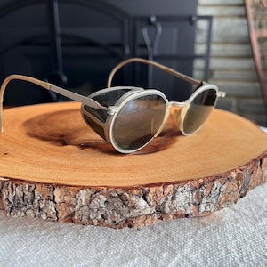 May include: Vintage style sunglasses with round lenses and side shields. The frames are silver-toned with gold-colored temples. The sunglasses are resting on a round, wooden slice with a natural bark edge.