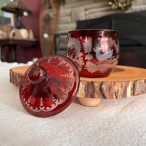 May include: A ruby red glass jar with a matching lid, both featuring etched white designs. The jar displays a forest scene with a deer. The lid is off to the side. The jar and lid are on a wooden slice.