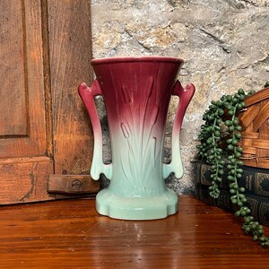 May include: A ceramic vase featuring a two-tone ombre glaze that transitions from a deep burgundy at the flared rim to a pale mint green at the base. The vessel has two symmetrical handles and a molded relief pattern of cattail plants.