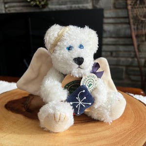 Pode incluir: Um urso de pel&uacute;cia branco com olhos azuis, nariz preto e aur&eacute;ola dourada. O urso tem asas bege e segura uma etiqueta com um desenho de floco de neve. O urso est&aacute; sentado em uma fatia de madeira.