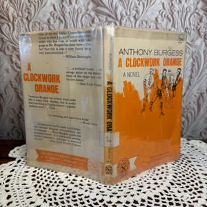 May include: A vintage copy of Anthony Burgess's novel "A Clockwork Orange." The book has an orange cover with white text and illustrations. The book is displayed open, revealing the title and author's name. The book is on a white lace doily.