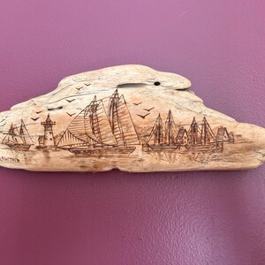 May include: A driftwood carving featuring a nautical scene with sailboats, a lighthouse, and birds. The artwork is etched into the light-colored wood, with the artist's signature visible. The piece is mounted on a purple background.