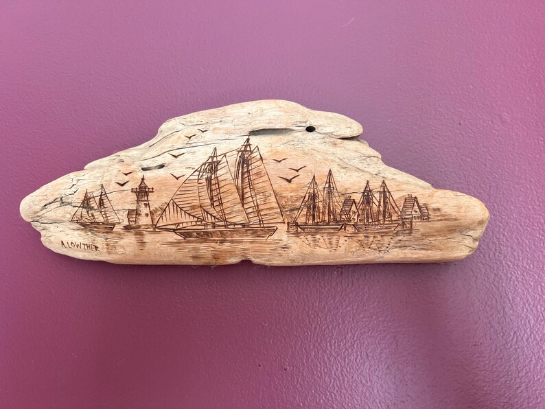 May include: A driftwood carving featuring a nautical scene with sailboats, a lighthouse, and birds. The artwork is etched into the light-colored wood, with the artist's signature visible. The piece is mounted on a purple background.