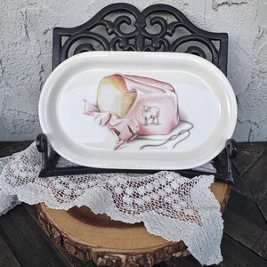 May include: An oval serving platter with a painted illustration of a loaf of bread wrapped in pink paper, tied with string, and a sheep illustration. The platter is white with a thin beige border and is displayed on a black metal stand.