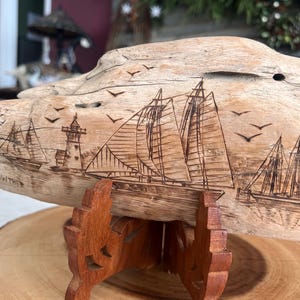 May include: A wooden sculpture of a nautical scene, featuring sailboats, a lighthouse, and flying birds. The artwork is carved into a piece of driftwood and mounted on a wooden stand. The sculpture is displayed on a wooden slice.