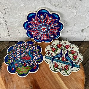 May include: Three decorative ceramic coasters with floral and bird designs. The coasters feature a blue, red, and white color scheme. One coaster has a peacock, another has two birds, and the third has a floral pattern.
