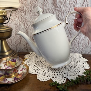 May include: White porcelain coffee pot with gold trim, held by a hand. A matching teacup and saucer with a fruit design sit on a wooden surface. A white lace doily and a brass lamp are also visible.