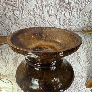 May include: A dark brown, glazed ceramic bowl with a wide, flared rim and a rounded, bulbous base. The surface features a mottled, glossy finish with subtle variations in tone, creating a reflective texture across the entire exterior.