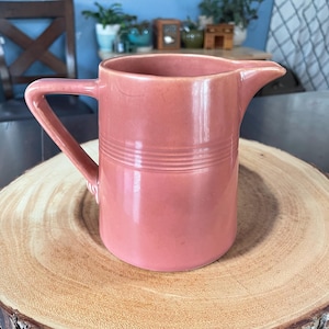 Puede incluir: Una jarra de cerámica rosa vintage con asa y boquilla. La jarra tiene detalles acanalados horizontales alrededor del centro y se encuentra sobre una rodaja de madera. El fondo incluye una pared azul y varias plantas.