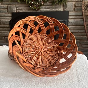 May include: A round, woven wicker basket with a decorative design. The basket is a warm brown color and features a smaller, woven insert in the center. The basket is sitting on a white surface.