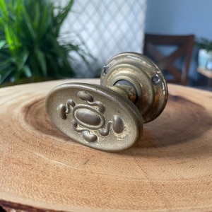 May include: An antique brass door knob with an oval handle and embossed design. The knob is set on a natural wood slice, highlighting its aged patina and intricate details. A vintage hardware piece, likely from the early 20th century.
