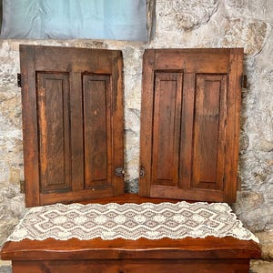 May include: Two rectangular wooden shutters with a dark brown finish and a two-panel design. Each shutter features vertical grain patterns, a recessed center, and metal hinges attached to the sides. The shutters are positioned upright.