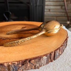 May include: A vintage brass nutcracker with a floral design on the handles, resting on a round wooden slab with a natural bark edge. The nutcracker is gold-colored and has a bulbous head.