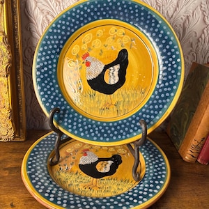 May include: Two ceramic plates featuring a chicken design. The plates have a yellow centre with a black and white chicken illustration, framed by a blue dotted border. The plates are displayed on a metal stand.