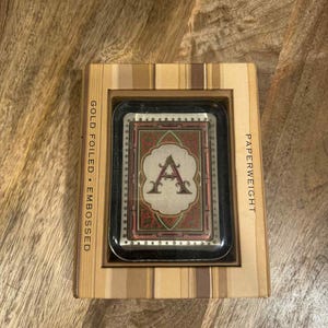 May include: A rectangular paperweight with the letter "A" in a decorative font, set within a frame. The paperweight is in a wooden box. The text "GOLD FOILED • EMBOSSED" and "PAPERWEIGHT" are printed on the box.