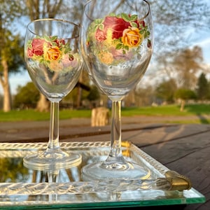 May include: Two clear wine glasses with gold rims and floral rose designs. The roses are red, yellow, and pink. The glasses sit on a mirrored tray with a gold trim. The background is a blurred outdoor scene with trees.