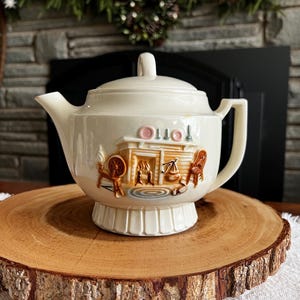 Puede incluir: Tetera de cerámica color crema con una escena decorativa que representa una chimenea, una rueca y una silla. La tetera tiene tapa y asa. La base es de diseño pedestal. La palabra "OTTO" está impresa en la tetera. La tetera se exhibe sobre una rodaja de madera.