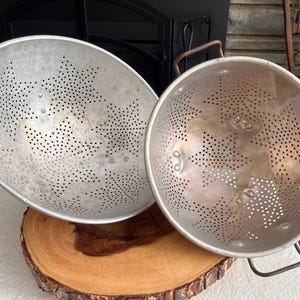 Op de afbeelding: Twee vintage metalen vergieten met stervormige perforatiepatronen. De vergieten zijn zilverkleurig en hebben handvatten. Ze rusten op een houten schijf, wat wijst op een rustiek of landelijk keukenthema.