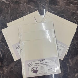 May include: Several cream-colored refill packs for a binder are displayed. Each pack is sealed in clear plastic and features a label with the text "Refill Pack-10" and "PVC Free". The packs are arranged on a dark, marbled surface.