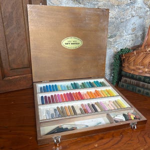 May include: A wooden box containing a collection of soft pastels arranged in three horizontal rows by color. The lid features an oval label reading "Talens Artists Soft Pastels". The set includes various shades of blue, pink, orange, and earth tones.