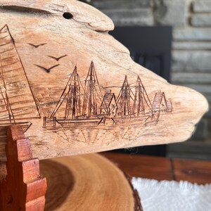 May include: A piece of driftwood with an etched nautical scene. The artwork depicts sailboats, buildings, and birds in flight. The wood has a natural, light brown color and is mounted on a wooden stand.