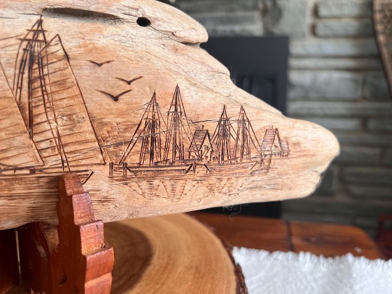 May include: A piece of driftwood with an etched nautical scene. The artwork depicts sailboats, buildings, and birds in flight. The wood has a natural, light brown color and is mounted on a wooden stand.