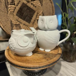 May include: A white ceramic owl-shaped tea set, including a teapot with a lid, a creamer, and a lidded sugar bowl. The set is displayed on a round wooden tray with a metal base. The background includes a woven basket and greenery.
