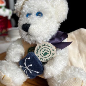 Pode incluir: Um urso de pel&uacute;cia branco com olhos azuis, um nariz preto e uma fita roxa escura. O urso tem uma etiqueta que diz "Boyds Bears Bearwear" e est&aacute; segurando uma pequena almofada bordada azul escura. O urso est&aacute; sentado em uma superf&iacute;cie de madeira.