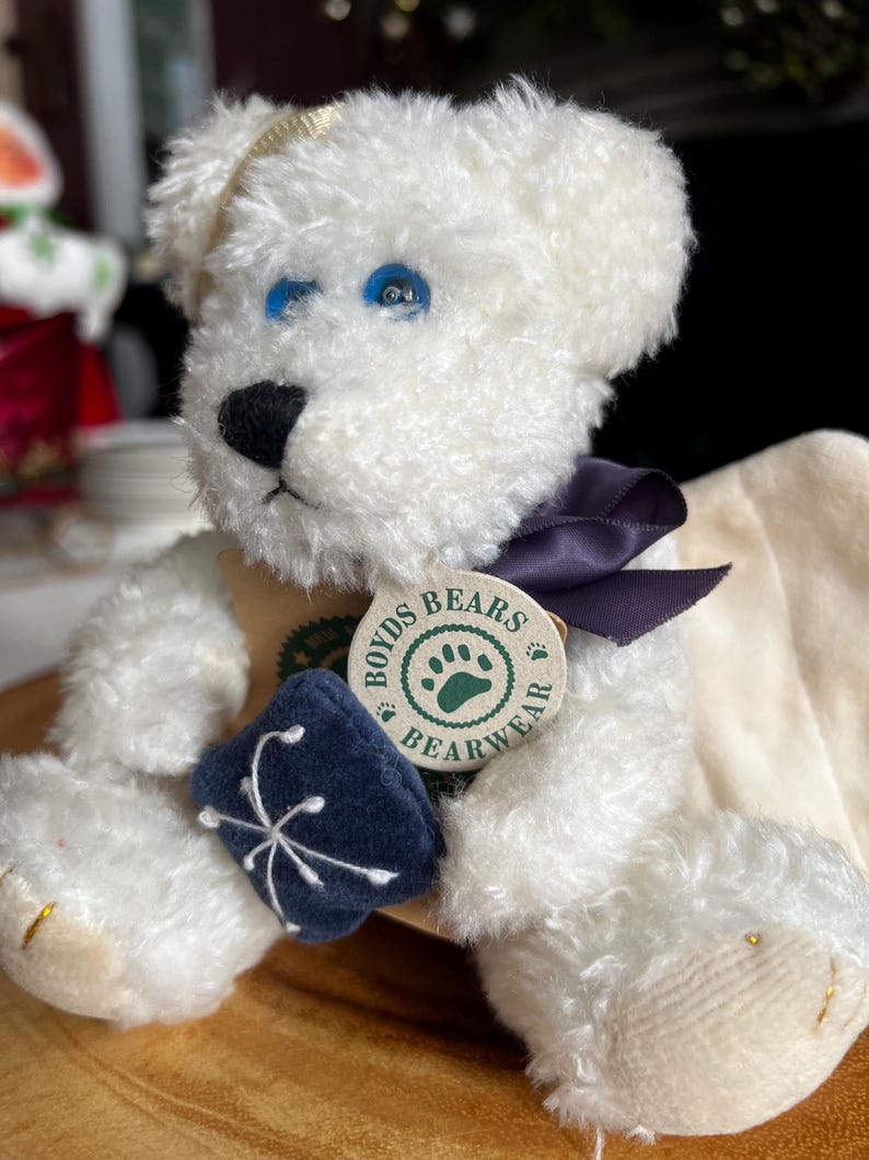 Pode incluir: Um urso de pel&uacute;cia branco com olhos azuis, um nariz preto e uma fita roxa escura. O urso tem uma etiqueta que diz "Boyds Bears Bearwear" e est&aacute; segurando uma pequena almofada bordada azul escura. O urso est&aacute; sentado em uma superf&iacute;cie de madeira.