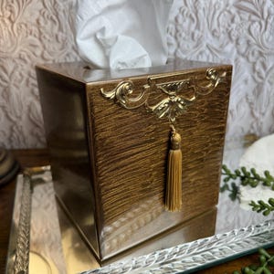 May include: A square tissue box cover with a metallic bronze finish and a textured surface. The front features a gold-toned ornate metal embellishment centered at the top, from which a matching gold-toned fabric tassel hangs vertically.
