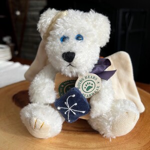 Pode incluir: Um urso de pel&uacute;cia branco com olhos azuis, nariz preto e uma faixa de cabe&ccedil;a dourada. O urso tem um quadrado azul escuro com um desenho de floco de neve branco e uma etiqueta que diz "Boyds Bears Bearwear". O urso est&aacute; sentado em uma superf&iacute;cie de madeira.