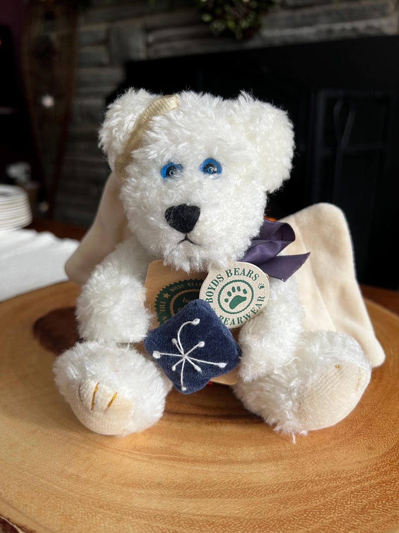 Pode incluir: Um urso de pel&uacute;cia branco com olhos azuis, nariz preto e uma faixa de cabe&ccedil;a dourada. O urso tem um quadrado azul escuro com um desenho de floco de neve branco e uma etiqueta que diz "Boyds Bears Bearwear". O urso est&aacute; sentado em uma superf&iacute;cie de madeira.