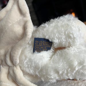 Pode incluir: Close-up de um urso de pel&uacute;cia creme. O urso tem uma textura macia e fofa e uma etiqueta com texto. A etiqueta diz "TBC LTD &copy;1990-98 ALL NEW MATERIALS CONTENTS: POLYESTER FIBER AND/OR PLASTIC BEADS HANDMADE".